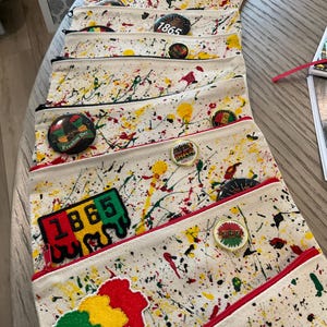 Puede incluir: Una colección de estuches de lona color hueso con cremallera y un patrón de salpicaduras de pintura multicolor. Cada estuche está decorado con varios pines circulares y parches de tela, incluyendo diseños con el texto 1865, Freedom Day y Love.