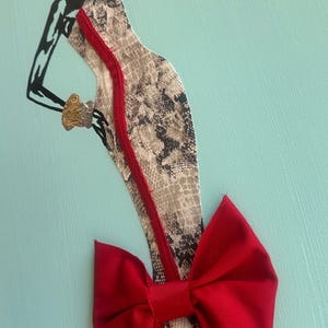 Puede incluir: Obra de arte de una figura estilizada con un vestido con estampado de piel de serpiente y un lazo rojo. La figura es en blanco y negro, con una pulsera dorada. El vestido tiene una cremallera roja sobre un fondo azul claro.