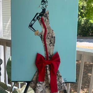 Puede incluir: Una obra de arte en lienzo que presenta una figura estilizada con un vestido con estampado de piel de serpiente y un gran lazo rojo. La figura tiene detalles en negro sobre un fondo azul claro. La obra se exhibe en un caballete negro.