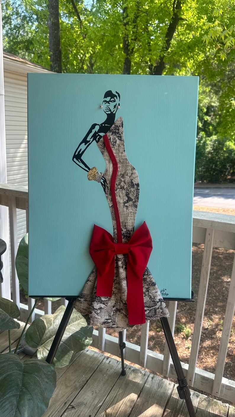 Puede incluir: Una obra de arte en lienzo que presenta una figura estilizada con un vestido con estampado de piel de serpiente y un gran lazo rojo. La figura tiene detalles en negro sobre un fondo azul claro. La obra se exhibe en un caballete negro.