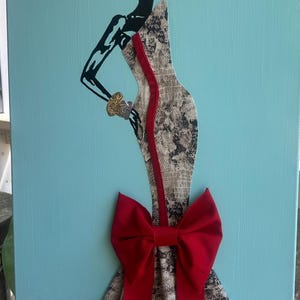 Puede incluir: Obra de arte en lienzo que presenta una silueta estilizada de una persona con un vestido con estampado de piel de serpiente y un gran lazo rojo. La figura tiene brazos negros y cabeza negra. Una raya roja recorre el centro del vestido. El fondo es azul claro.