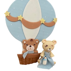 May include: A blue and white hot air balloon with a brown banner and yellow stars. Two teddy bears are in the basket, one brown and one white. The white bear is holding a blue blanket and a bottle.