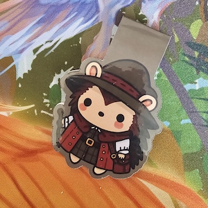Könnte beinhalten: Ein brauner und roter Comic-Igel, der einen Hut und einen Mantel mit einer Tasche trägt. Der Igel hält eine kleine Tasche in seiner Hand. Das Bild ist ein Lesezeichen.
