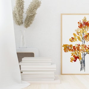 Op de afbeelding: Een ingelijst aquarel schilderij van een boom met oranje, gele en rode bladeren. De boom staat op een witte achtergrond en het frame is licht van kleur.