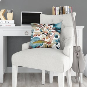 May include: A white desk and chair are in a room with a gray wall. The chair and a floor pillow have a watercolor floral design in shades of blue, brown, and green. A laptop, books, and a desk lamp are on the desk.
