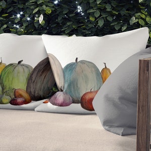 Może przedstawiać: Dwie białe poduszki dekoracyjne z akwarelą przedstawiającą różne dynie w odcieniach pomarańczu, zieleni i brązu. Poduszki leżą na jasnobeżowej sofie ogrodowej z drewnianą ramą, na tle zielonych liści.