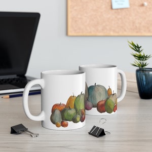 May include: Two white ceramic mugs with a watercolor illustration of various pumpkins in shades of orange, green, and blue. The mugs are on a light-colored wooden surface, with a laptop and office supplies in the background.