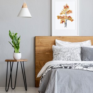 Op de afbeelding: Een slaapkamer met een houten hoofdeinde, een grijs dekbed en witte kussens. Een ingelijst aquarel schilderij van een boom met oranje en gele bladeren hangt boven het bed. Een klein nachtkastje met een plant staat naast het bed.