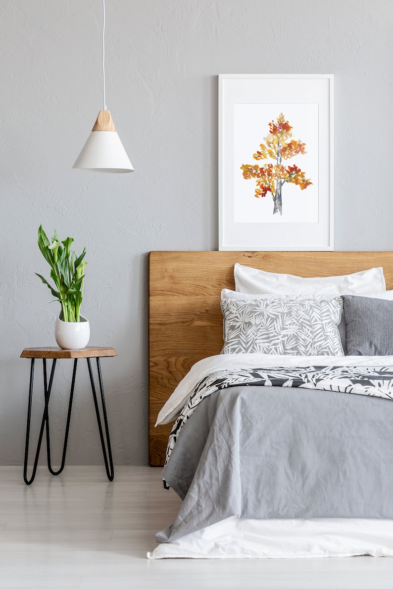 Op de afbeelding: Een slaapkamer met een houten hoofdeinde, een grijs dekbed en witte kussens. Een ingelijst aquarel schilderij van een boom met oranje en gele bladeren hangt boven het bed. Een klein nachtkastje met een plant staat naast het bed.