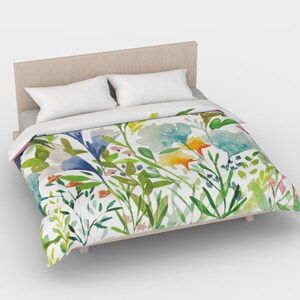 May include: A white duvet cover with a watercolor floral pattern featuring blue, green, yellow, and orange flowers. The duvet cover is on a bed with a wooden headboard and two white pillows.