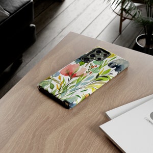 May include: A white phone case with a watercolor floral design featuring pink, yellow, green, and blue flowers. The case has a cutout for the camera lens.