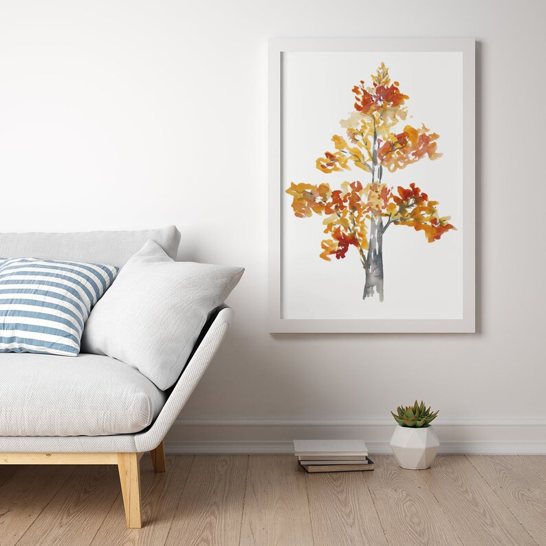 Op de afbeelding: Een aquarel van een boom met gele, oranje en rode bladeren. De boom is ingelijst in een wit frame en hangt aan een witte muur. Een witte bank met een blauw-wit gestreept kussen staat op de voorgrond.