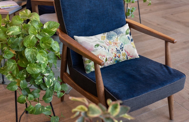 May include: A blue armchair with a floral patterned pillow. The chair has wooden arms and legs and is sitting on a wooden floor. A green plant is in the foreground.