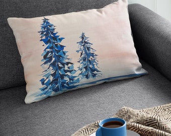 Puede incluir: Una almohada con un diseño de acuarela de dos pinos azules sobre un fondo rosa y blanco. La almohada está sobre un sofá gris con una taza de café azul y una manta.