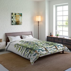 May include: A white duvet cover featuring a watercolour print of green and blue trees. The duvet cover is on a bed with white pillows and a brown headboard. The bed is in a bedroom with a white brick wall, a brown floor lamp, and a window with white blinds.