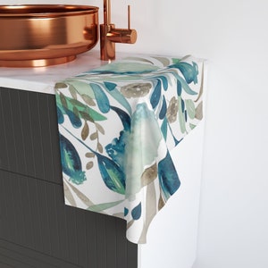 May include: A white hand towel with a watercolor floral pattern. The design features blue, teal, green, and brown leaves and flowers. The towel is draped over a white cabinet, near a copper sink and faucet.