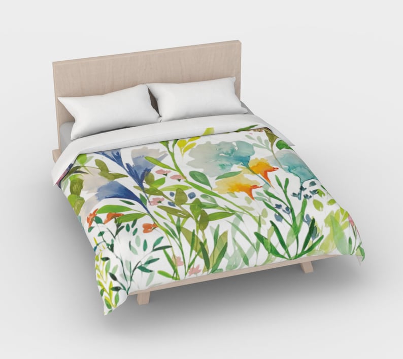 May include: A white duvet cover with a watercolor floral print featuring blue, yellow, green, and orange flowers. The duvet cover is on a bed with a wooden headboard and two white pillows.