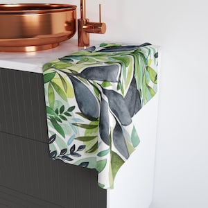 May include: A white cotton hand towel with a watercolour design of green and blue leaves. The towel is folded and placed on a countertop in a bathroom.