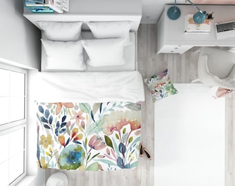 May include: A white bed with a floral duvet cover featuring watercolor flowers in shades of pink, blue, yellow, and green. The bed has two white pillows and a white throw pillow with a floral pattern.