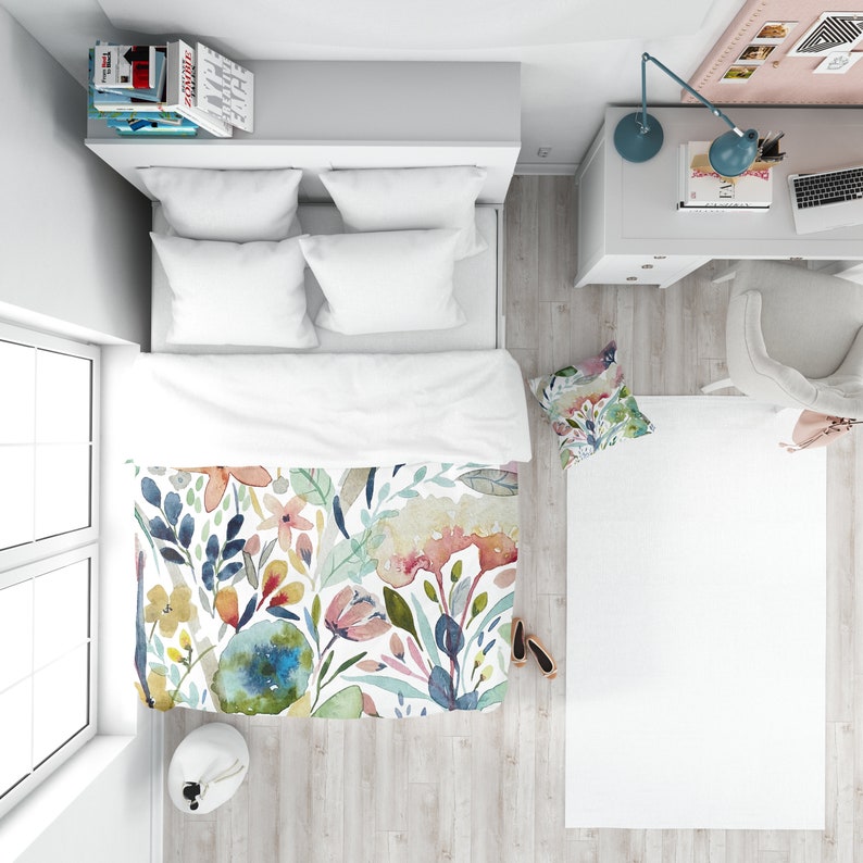 May include: A white bed with a floral duvet cover featuring watercolor flowers in shades of pink, blue, yellow, and green. The bed has two white pillows and a white throw pillow with a floral pattern.