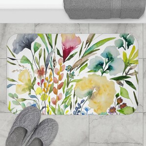 May include: A white bath mat with a watercolor floral design featuring yellow, blue, pink, and green flowers. The flowers are arranged in a bouquet-like pattern.
