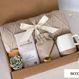 May include: A brown cardboard gift box filled with a cozy beige blanket, a mug, a small bag, a bar of chocolate, a small succulent plant, and a gold spoon. The box is labeled "BOX 1" and the chocolate bar is labeled "moonstruck, portland oregon, creamy milk chocolate, artisan chocolate".
