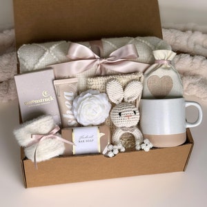 May include: A brown cardboard gift box filled with a variety of items, including a white blanket, a crocheted bunny, a white mug, a bar of soap, a white flower, and a small bag with a heart design. The box is tied with a pink ribbon.