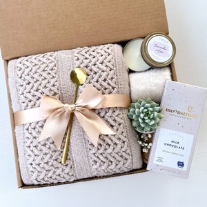 May include: A gift box filled with a cozy knitted throw blanket, a gold spoon, a candle with the label "lavender relax", a small succulent plant, and a bar of chocolate with the text "Creamy Milk Chocolate Artisan Chocolate".