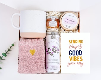 May include: A gift box filled with self-care items, including a white mug, pink fuzzy socks, a jar of honey, a tea bag, a candle, and a card that says "Sending Hugs & Good Vibes your way".