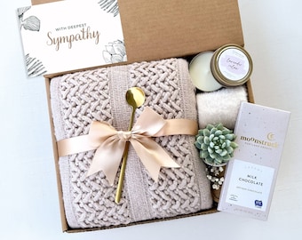 Può includere: Una scatola regalo piena di articoli per la cura di sé e il relax. La scatola include una morbida coperta di maglia rosa, un cucchiaio dorato, una candela, una piccola pianta grassa e una barretta di cioccolato al latte. La scatola è legata con un nastro di raso e include un biglietto che dice "Con le mie più sentite condoglianze".