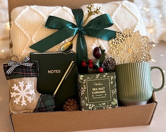 May include: A brown gift box filled with a variety of items, including a white blanket, a green mug, a notebook with the word "NOTES" on the cover, a small box of chocolate, a green and plaid stocking, and a gold snowflake ornament.