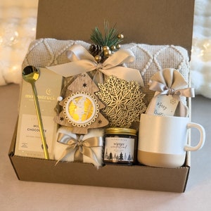 May include: A brown cardboard gift box filled with a cozy winter-themed gift set. The box contains a white blanket, a gold-colored metal snowflake ornament, a wooden Christmas tree ornament, a jar of candle, a mug, a bar of chocolate, and a gold spoon. The box is decorated with a gold ribbon and a tag that says "Merry Christmas".