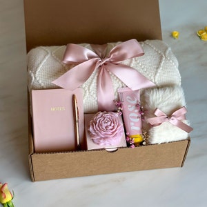 May include: A gift box filled with a white blanket, a pink notebook with a gold pen, a pink rose soap, a pink hand cream tube with the word "rose" on it, and a white fluffy item with a pink ribbon.