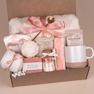 May include: A brown gift box filled with self-care items, including a white blanket, a pink bar of soap, a jar of bath salts, a white mug, a pink and white bath bomb, and a pink and white bath scrub. The box is tied with a pink ribbon.
