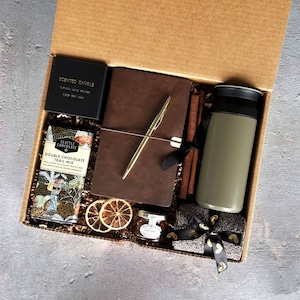 May include: A brown gift box filled with a variety of items, including a brown leather journal, a gold pen, a scented candle, a jar of jam, a bag of chocolate trail mix, a travel mug, and a grey scarf with gold ribbon.