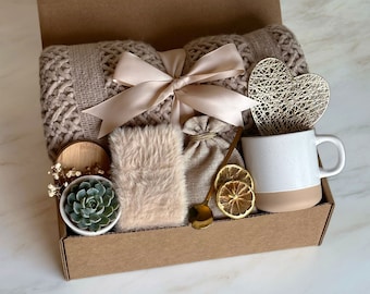 Puede incluir: Una caja de regalo de cartón marrón llena de una variedad de artículos, que incluyen una manta de punto, una pequeña suculenta en maceta, una taza, un objeto decorativo en forma de corazón y una pequeña bolsa. La caja está atada con una cinta beige.
