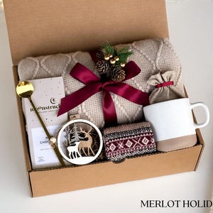 Op de afbeelding: Een bruine kartonnen cadeaudoos gevuld met een knusse winterse cadeauset. De doos bevat een beige gebreide deken, een mok, een paar sokken, een houten ornament met een hertontwerp, een chocolade reep en een gouden lepel. De doos is vastgebonden met een bordeauxrood lint en versierd met een kleine dennenappel en bessen.