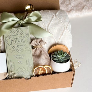 Peut inclure: Une bo&icirc;te-cadeau remplie d'une couverture verte et blanche, une tablette de chocolat Ritual avec le texte "Fleur de Sel Chocolat Noir avec Sel de Mer 70% Cacao", un petit sac en toile de jute, un pot en c&eacute;ramique blanc avec une plante succulente et des tranches de citron s&eacute;ch&eacute;es.