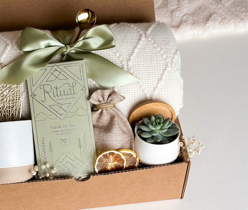 Peut inclure: Une bo&icirc;te-cadeau remplie d'une couverture verte et blanche, une tablette de chocolat Ritual avec le texte "Fleur de Sel Chocolat Noir avec Sel de Mer 70% Cacao", un petit sac en toile de jute, un pot en c&eacute;ramique blanc avec une plante succulente et des tranches de citron s&eacute;ch&eacute;es.