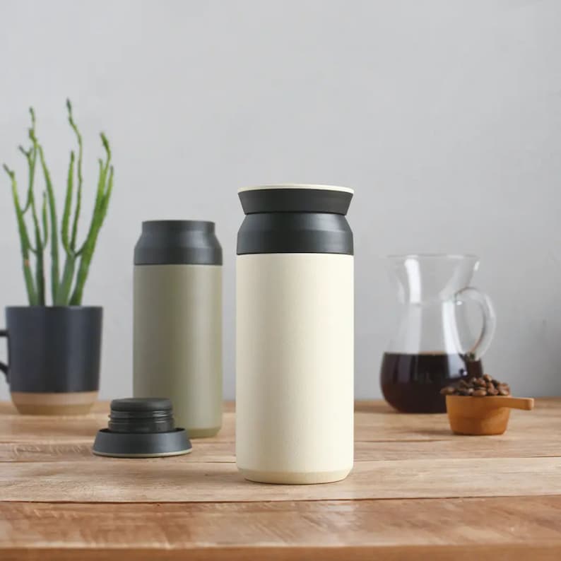Puede incluir: Taza de viaje aislada de color crema con tapa negra, junto a una taza verde y una jarra de vidrio transparente. La taza es cil&iacute;ndrica y parece estar hecha de acero inoxidable. Una peque&ntilde;a cuchara de madera con granos de caf&eacute; est&aacute; sobre la mesa de madera.
