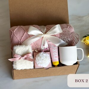 May include: A pink and white gift box filled with items for self-care, including a pink knitted throw blanket, a white mug, a white fluffy item, a pink and white box, and a yellow tube with the word "rose" on it.