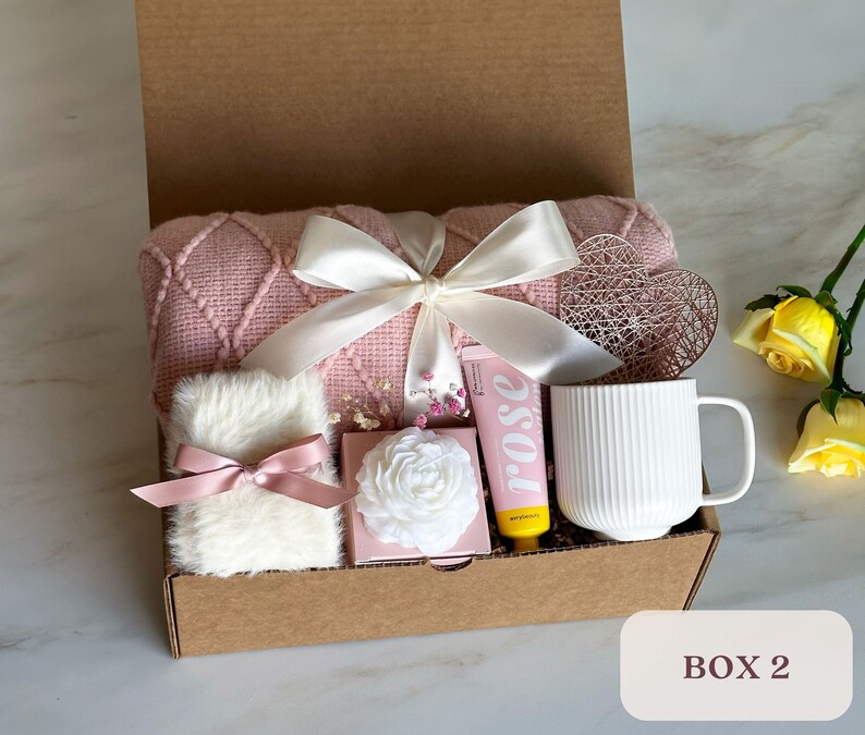 May include: A pink and white gift box filled with items for self-care, including a pink knitted throw blanket, a white mug, a white fluffy item, a pink and white box, and a yellow tube with the word "rose" on it.
