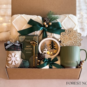 Op de afbeelding: Een cadeaupakket met een wit deken, een groene mok, een houten ornament met een hert en een kerstboom, een doos met warme chocolademelk, een groene en geruite sok met een sneeuwvlok en een gouden lepel. De mand is versierd met groen lint en een dennentak. De tekst "FOREST NOEL" staat op de bodem van de mand.