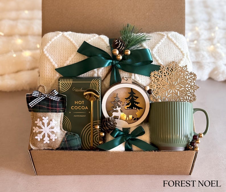 Op de afbeelding: Een cadeaupakket met een wit deken, een groene mok, een houten ornament met een hert en een kerstboom, een doos met warme chocolademelk, een groene en geruite sok met een sneeuwvlok en een gouden lepel. De mand is versierd met groen lint en een dennentak. De tekst "FOREST NOEL" staat op de bodem van de mand.