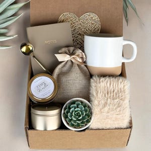 K&ouml;nnte beinhalten: Eine Geschenkbox gef&uuml;llt mit Selbstpflegeartikeln, darunter eine Tasse, eine Kerze, eine kleine Sukkulente, ein Notizbuch, ein L&ouml;ffel und ein Kunstfell. Die Box ist braun und hat einen goldenen Herz-Ausschnitt auf dem Deckel.