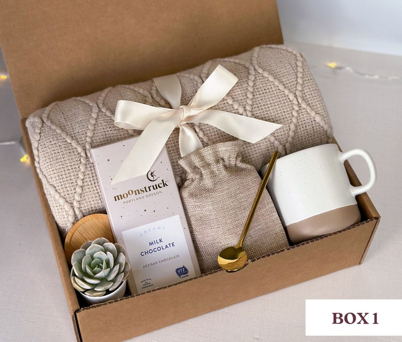 May include: A brown cardboard gift box filled with a cozy beige blanket, a mug, a small bag, a bar of chocolate, a small succulent plant, and a gold spoon. The box is labeled "BOX 1" and the chocolate bar is labeled "moonstruck, portland oregon, creamy milk chocolate, artisan chocolate".