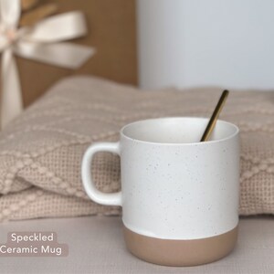 May include: A white ceramic mug with a speckled design and a brown bottom. The mug is sitting on a beige knitted blanket.