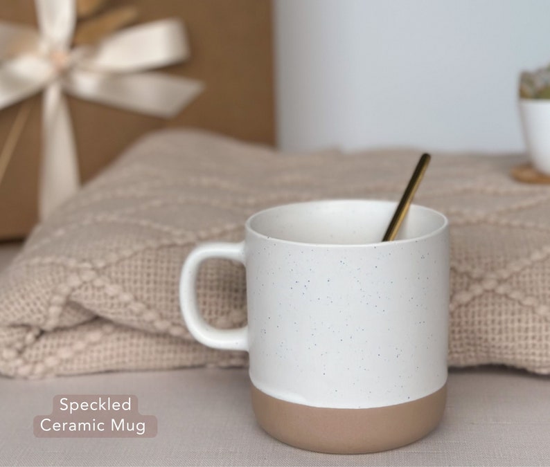 May include: A white ceramic mug with a speckled design and a brown bottom. The mug is sitting on a beige knitted blanket.