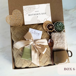 May include: A gift box filled with a brown throw blanket, a bar of chocolate, a wooden spoon, a mug, a small potted succulent, dried citrus slices, a heart-shaped decorative piece, and a card that says "Happy Mother's Day".