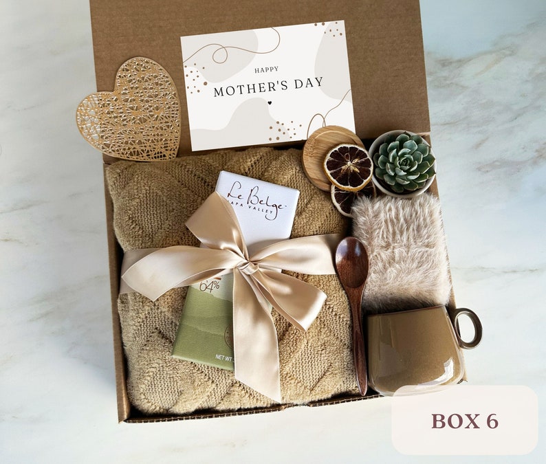 May include: A gift box filled with a brown throw blanket, a bar of chocolate, a wooden spoon, a mug, a small potted succulent, dried citrus slices, a heart-shaped decorative piece, and a card that says "Happy Mother's Day".
